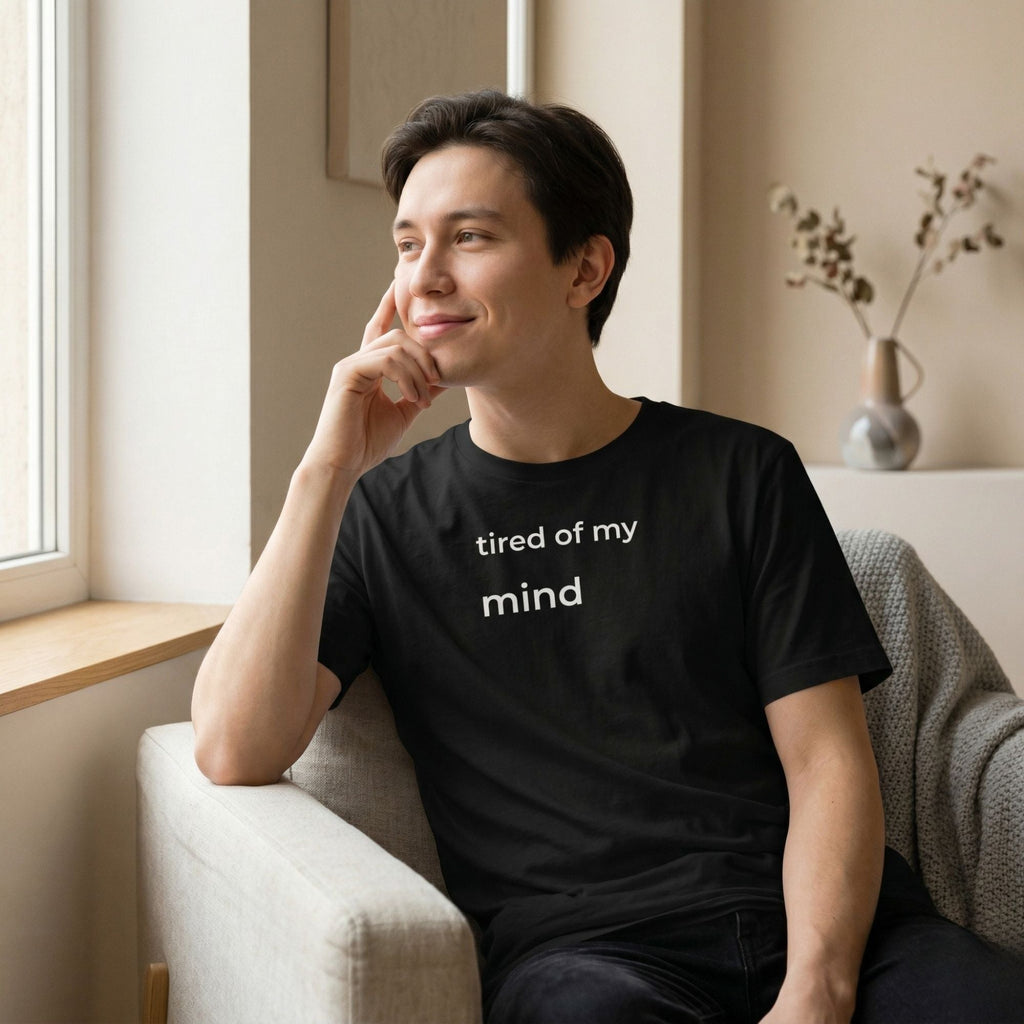 Man wearing black t-shirt with small text “tired of my mind”, sitting by window, natural light, relaxed minimal lifestyle scene