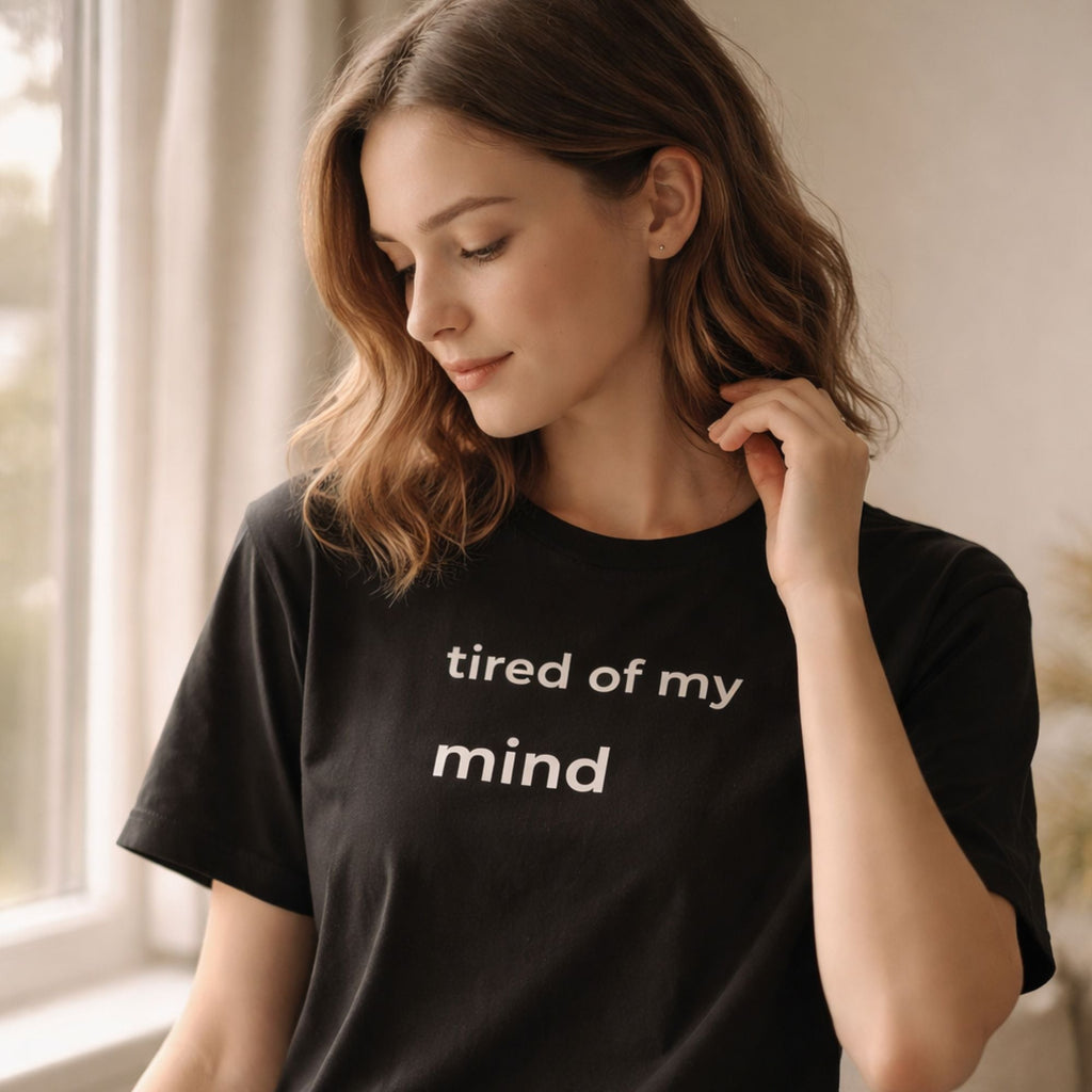 Woman wearing black minimalist t-shirt with text “tired of my mind”, soft natural light, calm indoor setting, introspective mood