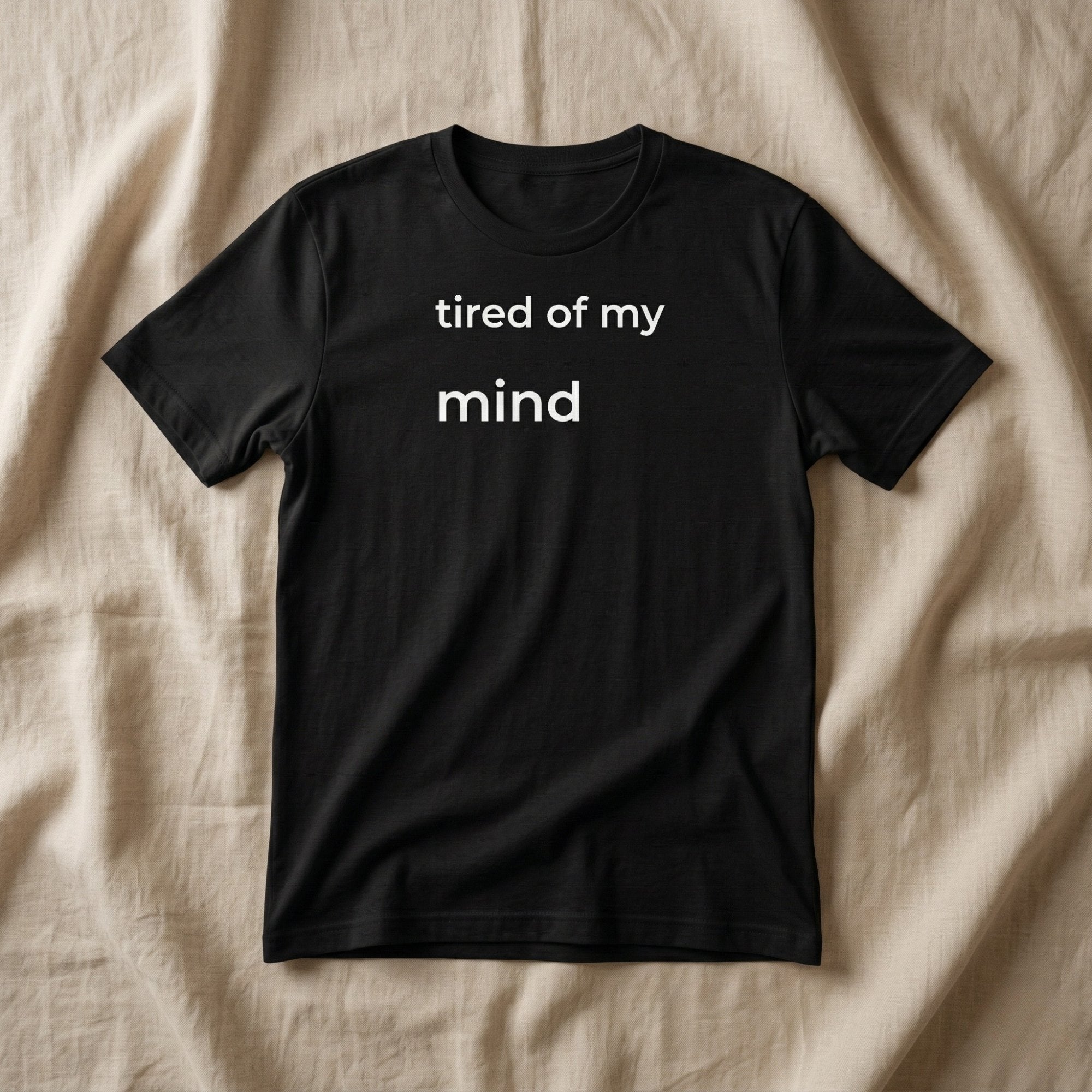Black minimalist t-shirt with small white text “tired of my mind” laid flat on neutral fabric, soft natural light, clean aesthetic